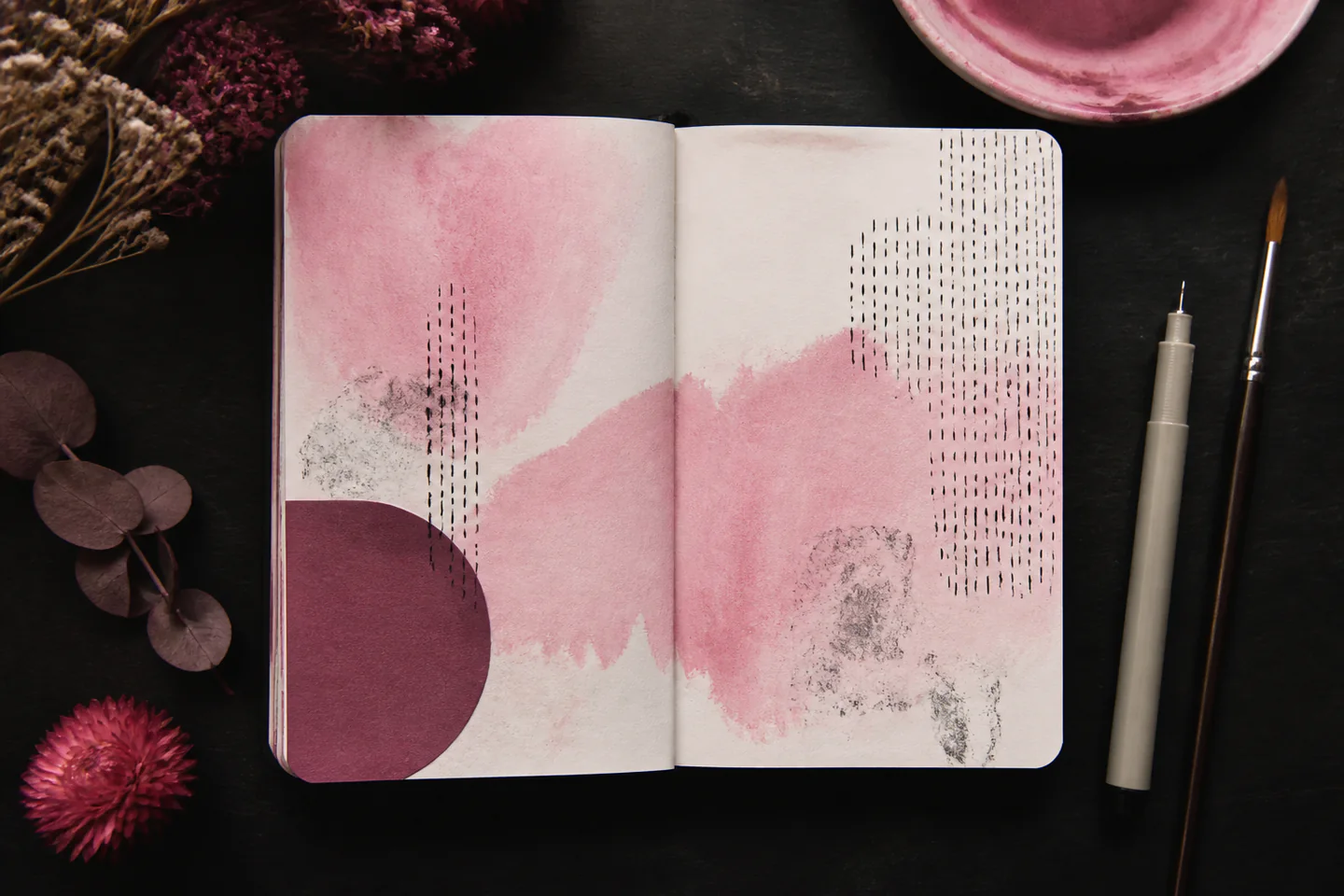 An open mixed-media art journal with a painted colour block, ink lines and one small collage beside pens and a brush