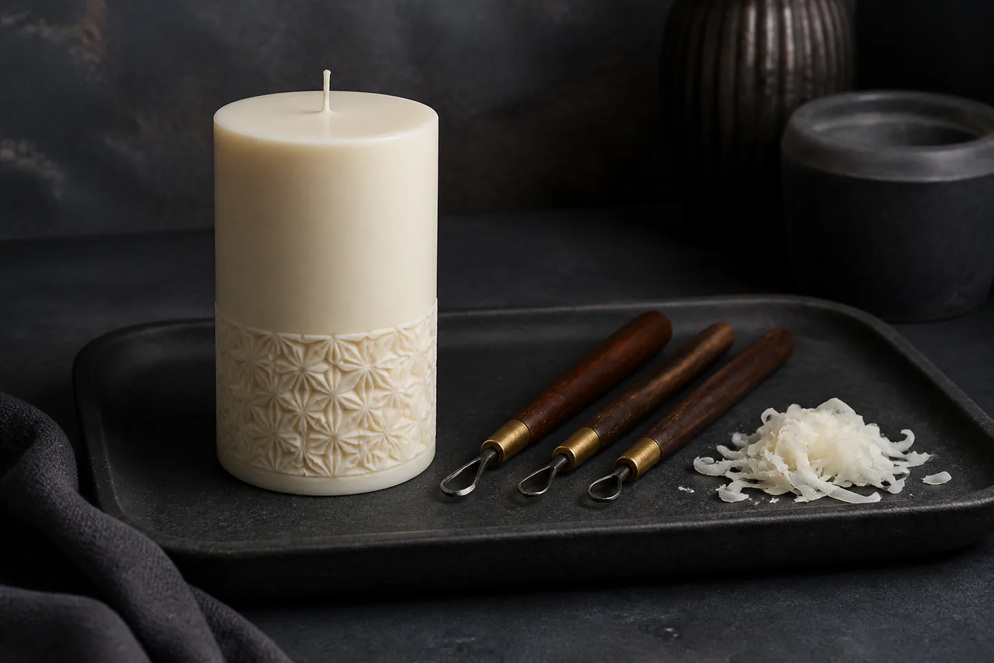 Unlit ivory pillar candle with shallow geometric carved bands beside small wax carving tools and collected wax shavings on a dark heatproof table with no people, flame, text or logos