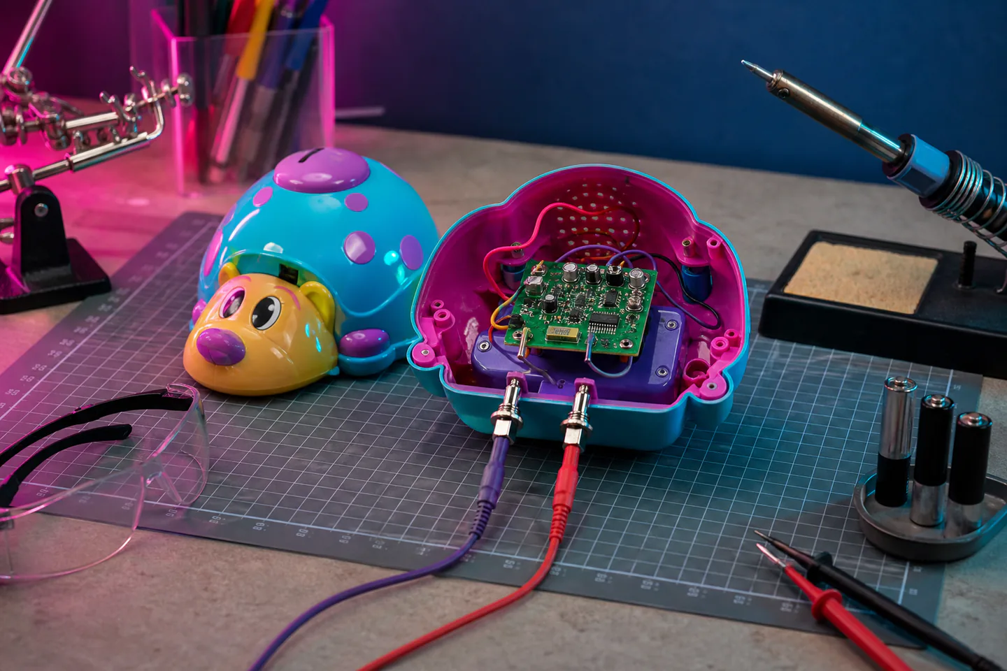 Opened battery-powered sound toy with AA holder, speaker, circuit board, insulated clip leads and small switches on a clear electronics bench