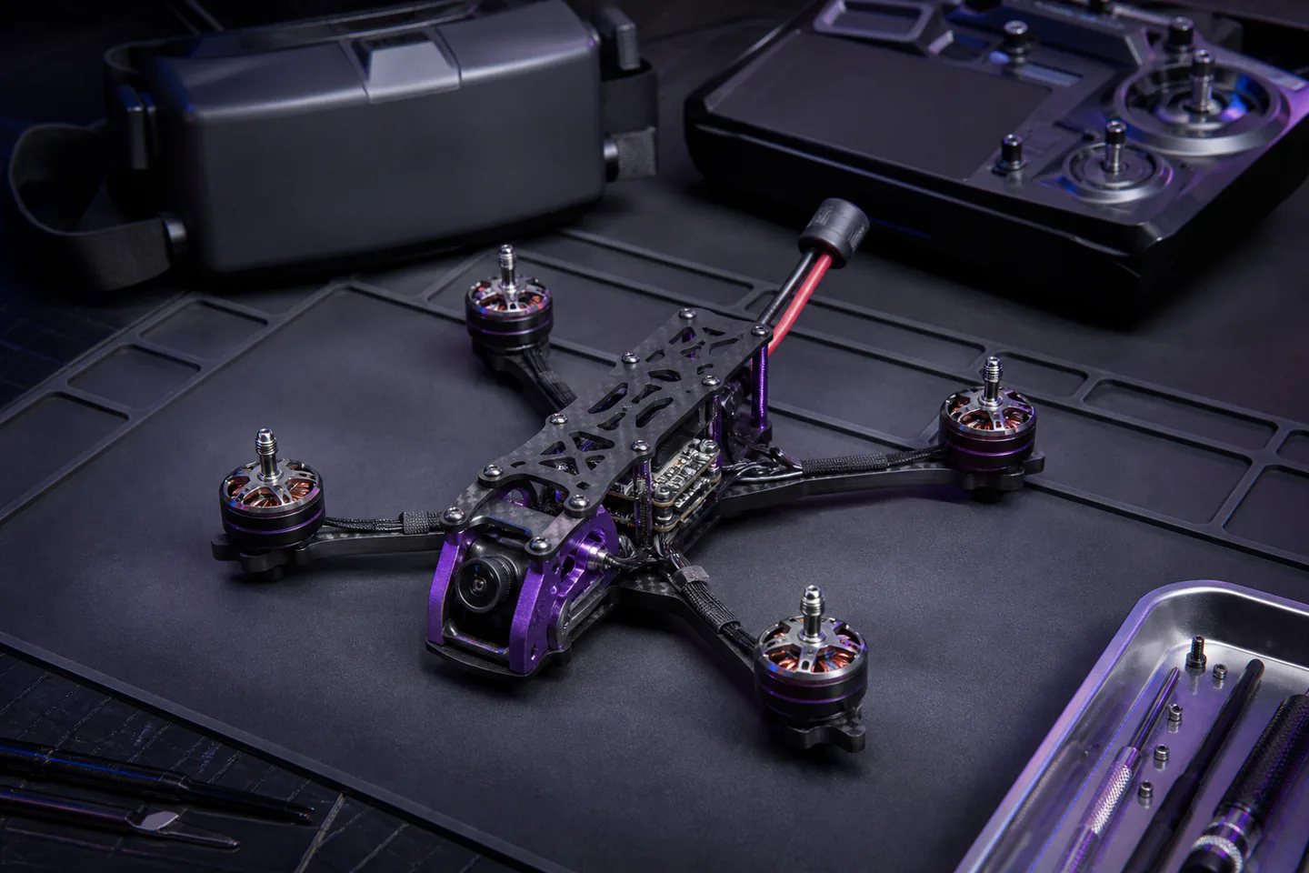 Small ducted FPV quadcopter beside goggles, radio controller and batteries, with a larger unbuilt carbon frame in the background and no hands or text