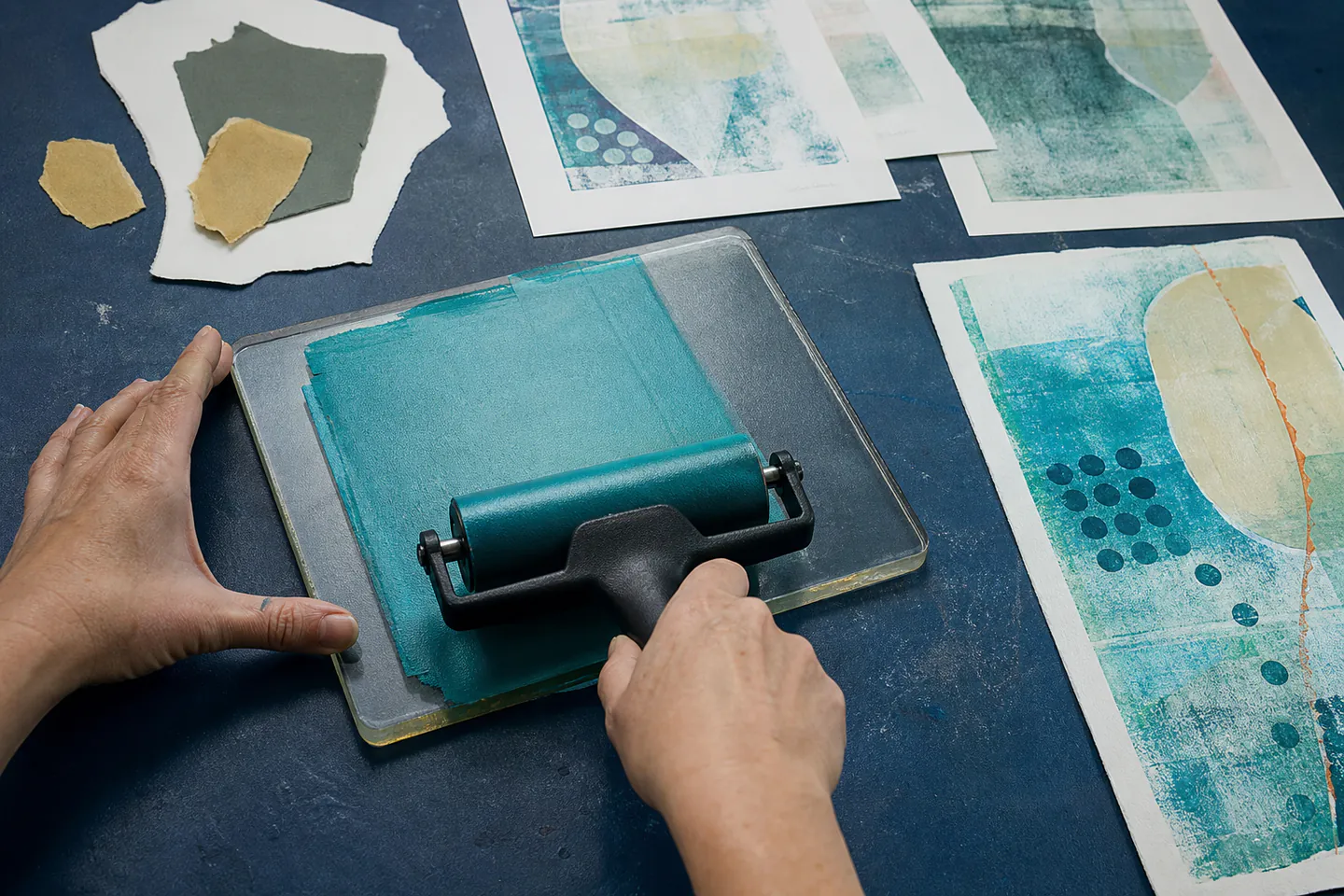 Hands rolling a thin blue acrylic layer across a rectangular gel printing plate beside paper masks and fresh monoprints