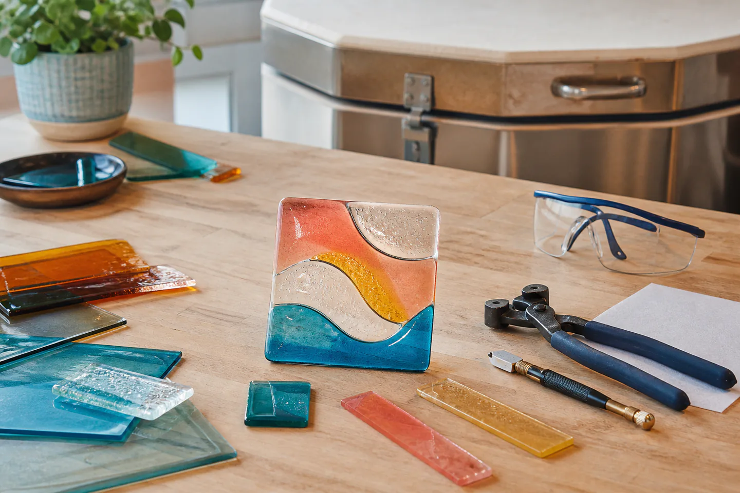 Small colourful fused-glass tile beside compatible labelled glass pieces, kiln paper, cutter and protective glasses on a studio bench