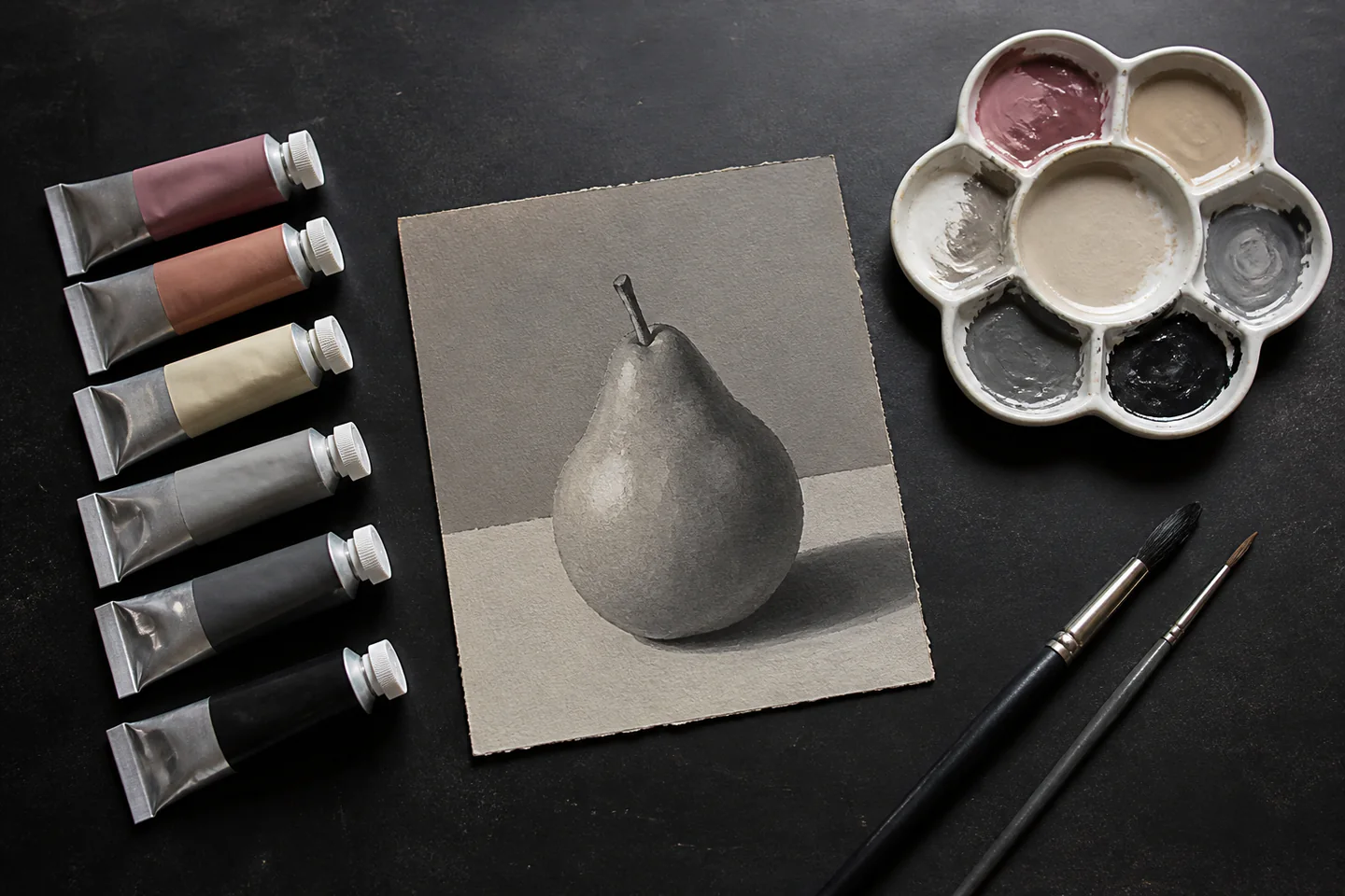 A small set of gouache paint tubes beside a matte painted pear study, two brushes and a ceramic palette on a dark studio table, no people, text or logos