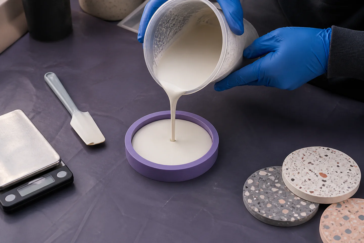 Gloved hands pouring pale speckled Jesmonite mixture into a simple round silicone coaster mould on a protected workbench