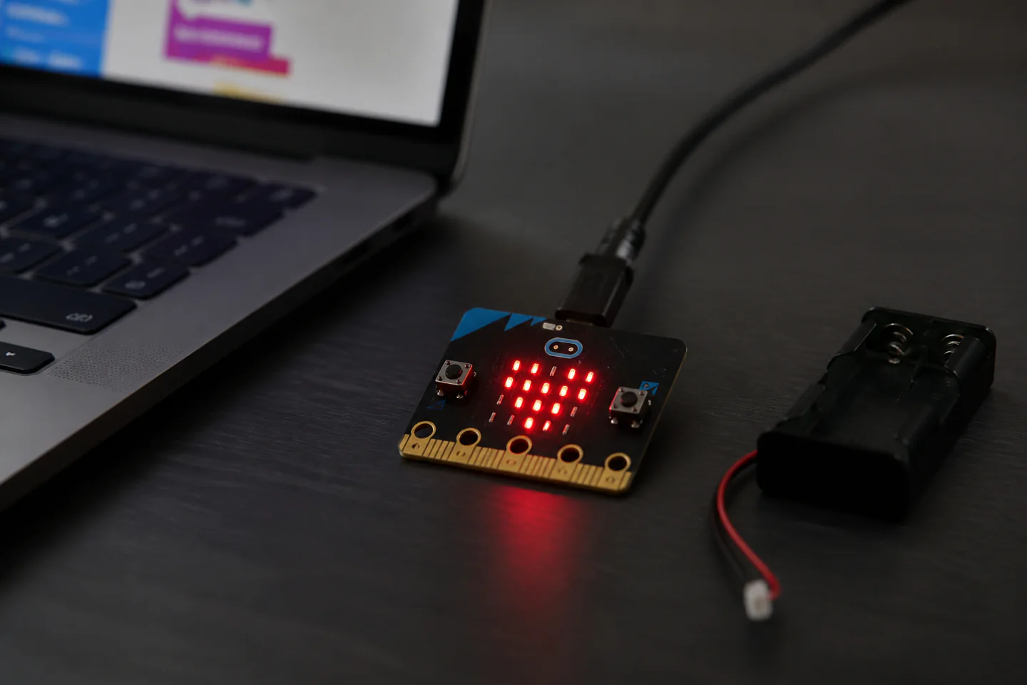A BBC micro bit board connected by USB showing a simple LED heart beside a laptop with block code and a few safely stored components