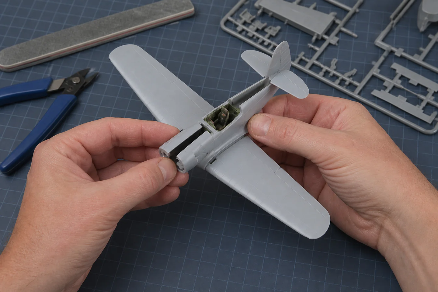 Hands aligning the wings and fuselage of a small unpainted plastic Spitfire model over a cutting mat and instruction sheet