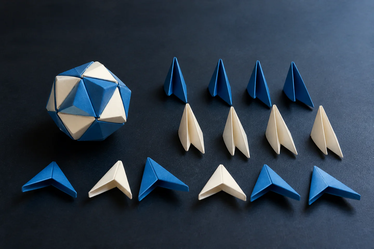 Several precisely folded blue and cream modular origami units beside a partly assembled geometric form on a dark table