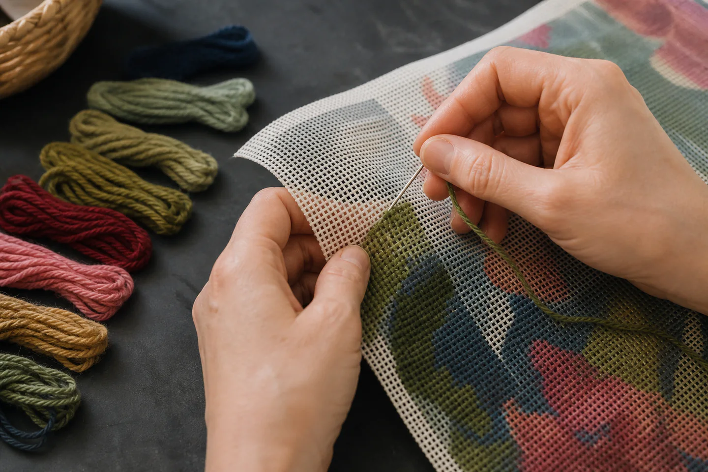 Hands stitching coloured wool through a printed needlepoint canvas beside organised yarn and a blunt tapestry needle