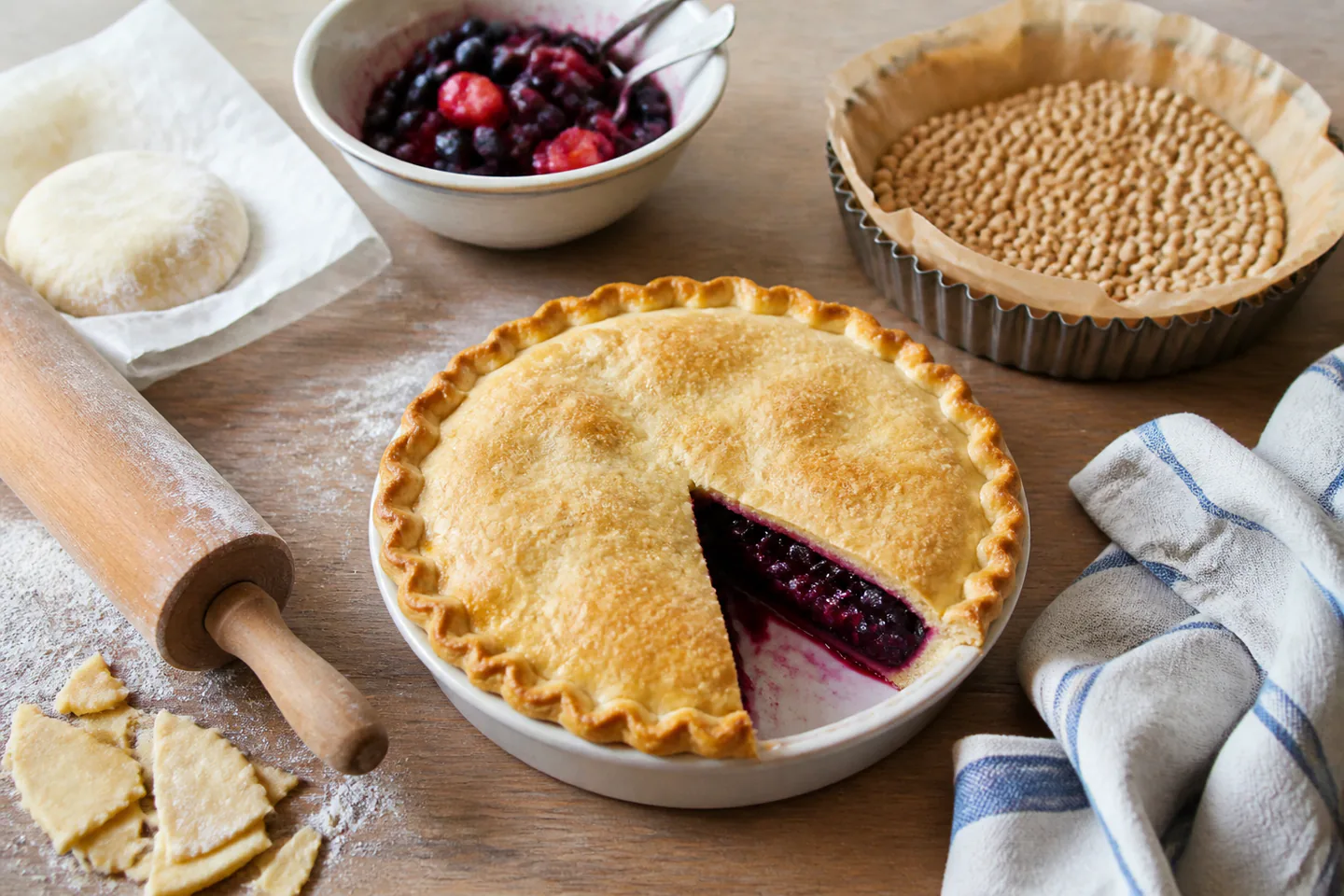 Golden fruit pie in a ceramic dish beside a blind-baked pastry case, rolling pin, pastry offcuts and a bowl of fruit filling