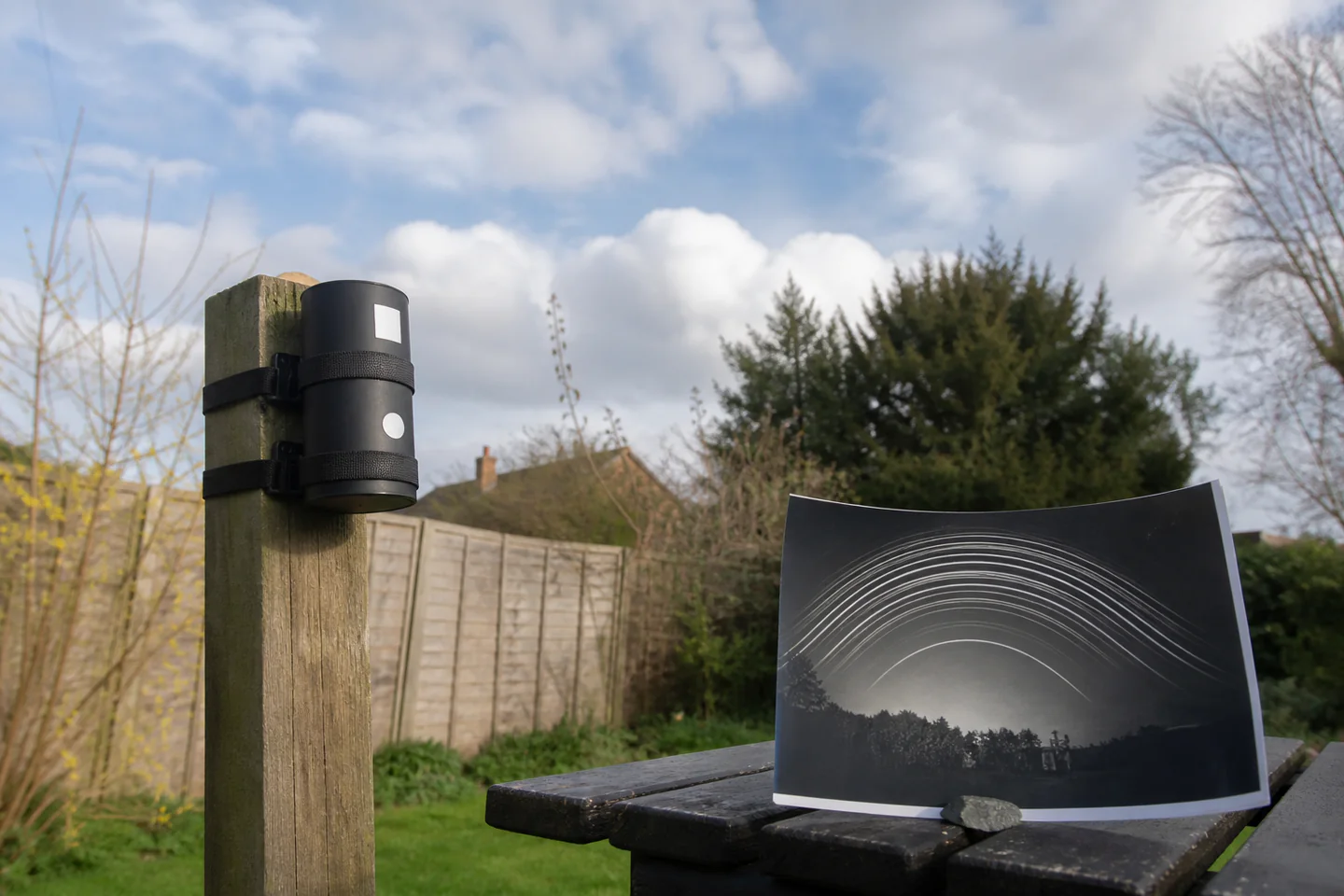 A small weatherproof pinhole can camera secured to a garden post beneath an open sky, with a developed solargraph showing curved sun trails beside it, no text or logos
