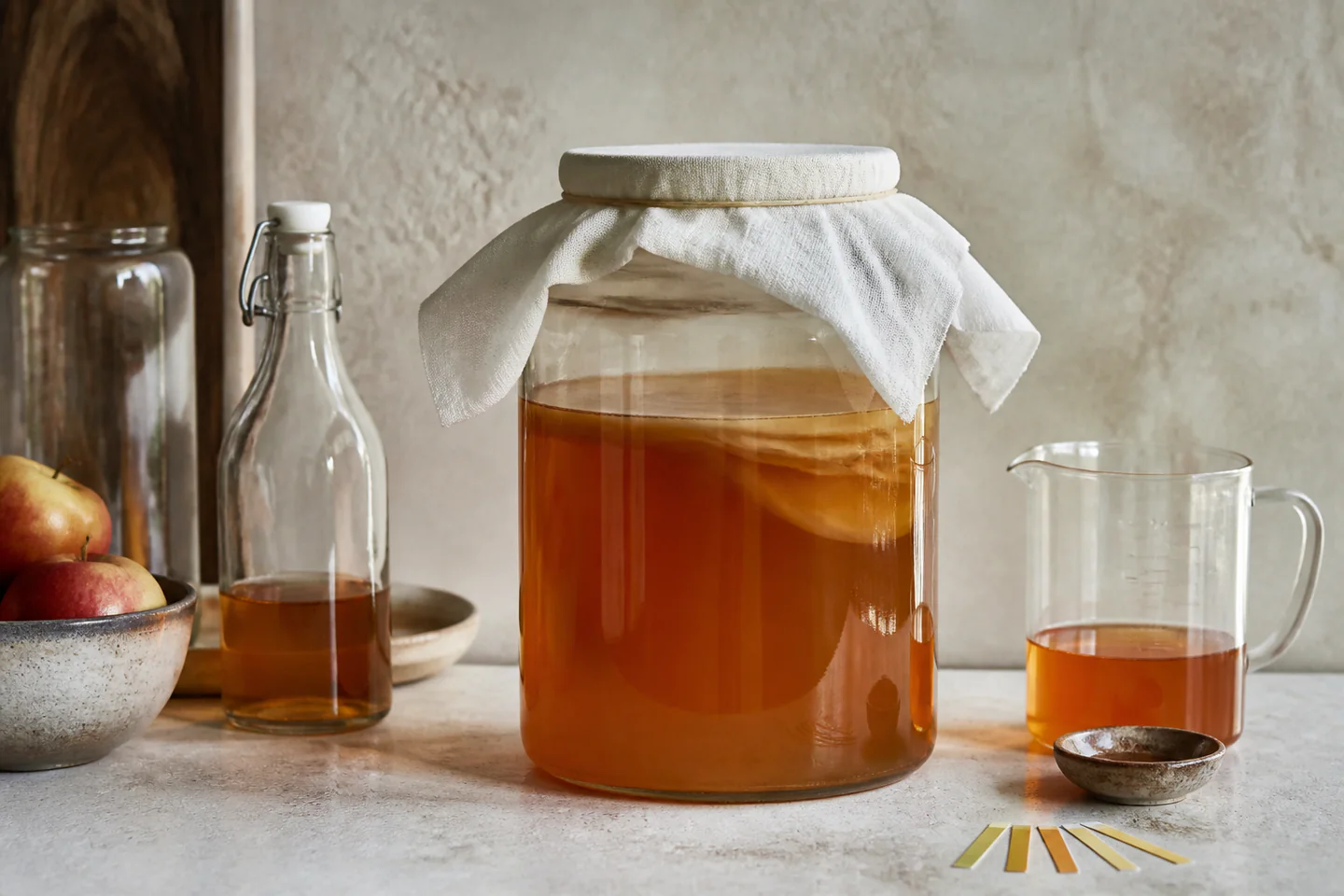 Wide glass vinegar jar covered with cloth beside a small bottle of cider, pH strips and a clean spoon